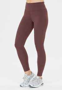Mörkröda högmidjade leggings gjorda av elastiskt tyg. Har en jämn textur och förstärkta sömmar. Kombineras med grå sportskor.