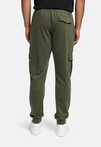 Man in olijfgroene joggerbroek met cargozakken en witte sneakers, staand met zijn rug naar de camera tegen een effen achtergrond.