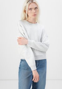 Felpa grigia dal taglio rilassato, con polsini e orlo a costine, texture morbida e spalle scese, abbinata a jeans azzurri chiari.