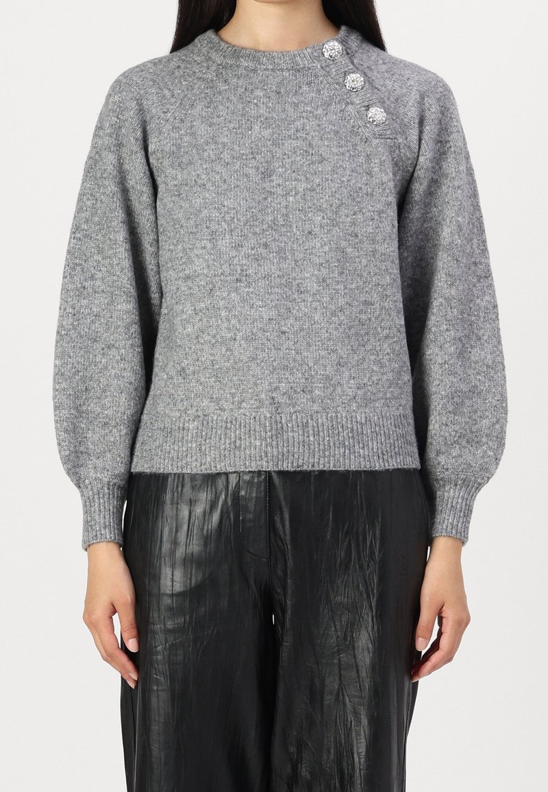 Pull en maille gris avec trois boutons décoratifs sur l'épaule, associé à un pantalon noir ample texturé, porté par une personne debout.