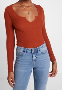 Body a maniche lunghe, color ruggine, con scollatura a V profonda e trama a costine, abbinato a jeans a vita alta di un blu chiaro. Collana a catena dorata come accento.