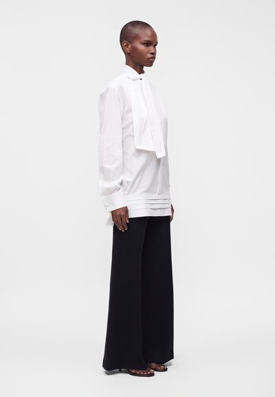 Personne aux cheveux courts portant un chemisier blanc à manches longues avec un nœud devant et un pantalon noir à jambes larges, debout de profil.
