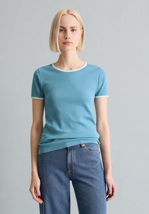 Femme aux cheveux blonds courts portant une chemise bleu clair à manches courtes avec bordure blanche et un jean bleu taille haute sur un fond uni.