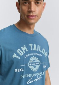 Modrá bavlnená tričko s bielym grafickým potlačou "TOM TAILOR" a detailami okrúhleho loga. Klasický okrúhly výstrih, krátke rukávy, hladká textúra.