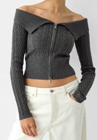 Gray off-shoulder sweater with ribbed texture and cable-knit design, featuring a front zipper and cropped fit. Paired with white high-waisted pants.