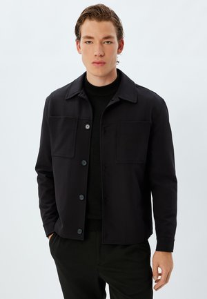 Black jacket made of smooth fabric, featuring a collar, two front pockets, and button closure. Worn over a black sweater and trousers.
