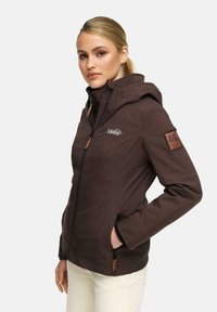 Vrouw met blond haar die een bruine hoodiejas met ritsen en patches draagt, staande met één hand in de zak tegen een effen achtergrond.