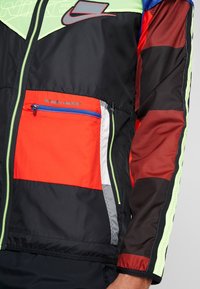 Veste avec des sections noires, rouges, vert néon et bleues, dotée d'une fermeture éclair et d'une poche rouge portant l'inscription « PACKABLE GARMENT ».