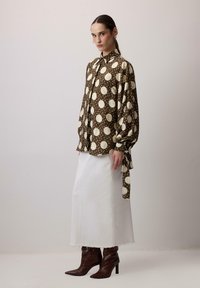 Blusa marrone a pois con maniche ampie, abbinata a una gonna di jeans bianca e stivali alti marroni, che mostrano una silhouette moderna.