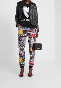 Giacca da motociclista in pelle nera, t-shirt nera con grafica di un cuore argentato, pantaloni a fantasia con cassette colorate, borsa nera e décolleté bicolor.