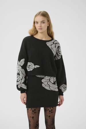 Maglione nero con grande ricamo di rosa grigia sulle maniche e sul davanti, abbinato a una gonna coordinata. Il modello indossa collant con motivo floreale.