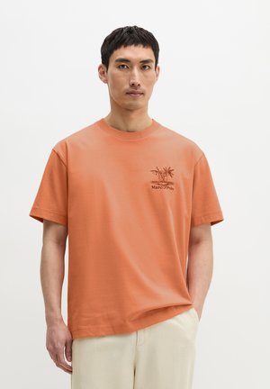 Mann trägt ein schlichtes orangenes Kurzarm-T-Shirt mit einer kleinen Pflanzengrafik und Text auf der linken Brustseite, kombiniert mit hellbeigen Hosen.