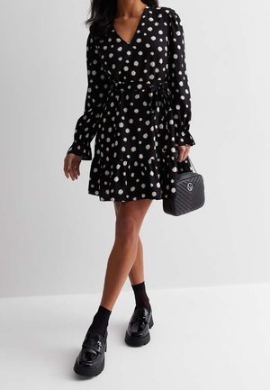 Czarna sukienka w kropki z dekoltem w serek, długimi rękawami i paskiem w talii, zestawiona z czarnymi butami na platformie oraz torebką o fakturze.