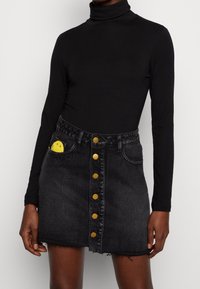 Maglione a collo alto nero abbinato a una gonna di denim nera con bottoni dorati e un sorriso ricamato giallo sulla tasca anteriore.