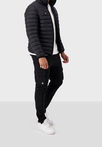 Giubbotto imbottito nero con cuciture orizzontali, chiusura con zip e tasche laterali, abbinato a pantaloni cargo neri con tasche a patta e sneakers bianche.