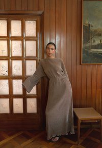 Robe longue marron texturée avec des manches évasées, taille cintrée. Le mannequin s'appuie contre une porte en bois avec des panneaux dépoli, le sol en bois est visible.