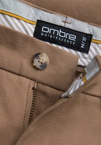 Pantalon beige avec fermeture éclair ouverte, boutonnière fermée, et étiquette intérieure visible portant la mention "ombre Quintessence M/32" sur la ceinture rayée.