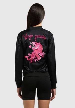 Nő fekete szatén bomber dzsekiben, melynek hátulján élénk rózsaszín hímzett tigris és a "kujo genuine" felirat látható, fekete rövidnadrággal párosítva.