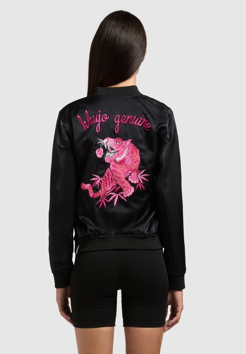 Femeie purtând o jachetă bomber din satin negru cu tigru brodat roz aprins și textul „kujo genuine” pe spate, asortată cu pantaloni scurți negri.