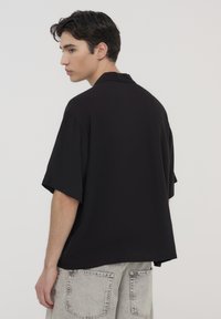 Camicia nera a maniche corte con una vestibilità rilassata e colletto, realizzata in tessuto morbido, indossata con pantaloni corti in denim grigio chiaro con grandi tasche posteriori.