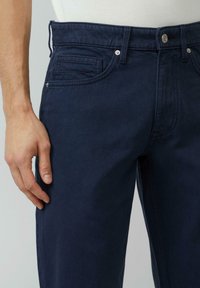 Granatowe dżinsy mają gładką teksturę, prosty krój nogawek, przednie kieszenie oraz metalowe akcenty przy pasie.