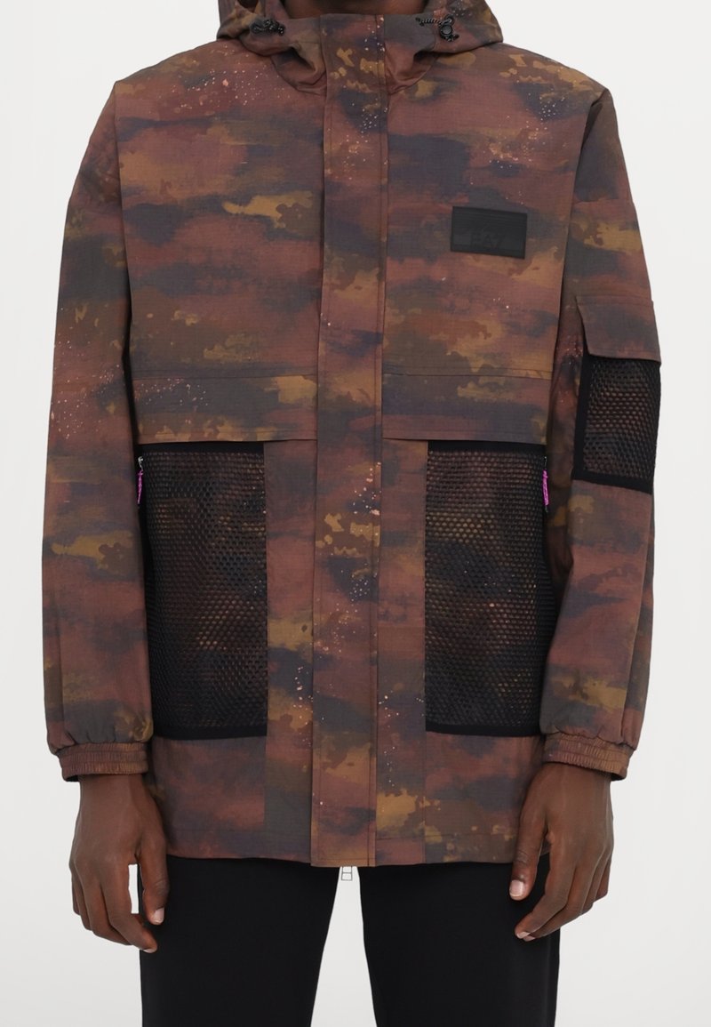 Veste camouflage marron et noire avec poches en maille noire sur le devant et la manche, fermeture éclair à l'avant, capuche, portée par une personne à la peau foncée.