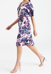Sukienka kopertowa w kwiaty w kolorze białym z fioletowo-niebieskimi wzorami, z talią podkreśloną paskiem, sięgająca kolan, z krótkimi rękawami. Do tego lawendowe sandały na obcasie z paskami.