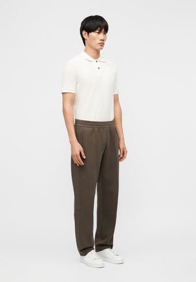 Νεαρός άνδρας στέκεται φορώντας λευκό πόλο με κοντά μανίκια, καφέ παντελόνι χαλαρής εφαρμογής και λευκά αθλητικά παπούτσια, μπροστά σε απλό φόντο.