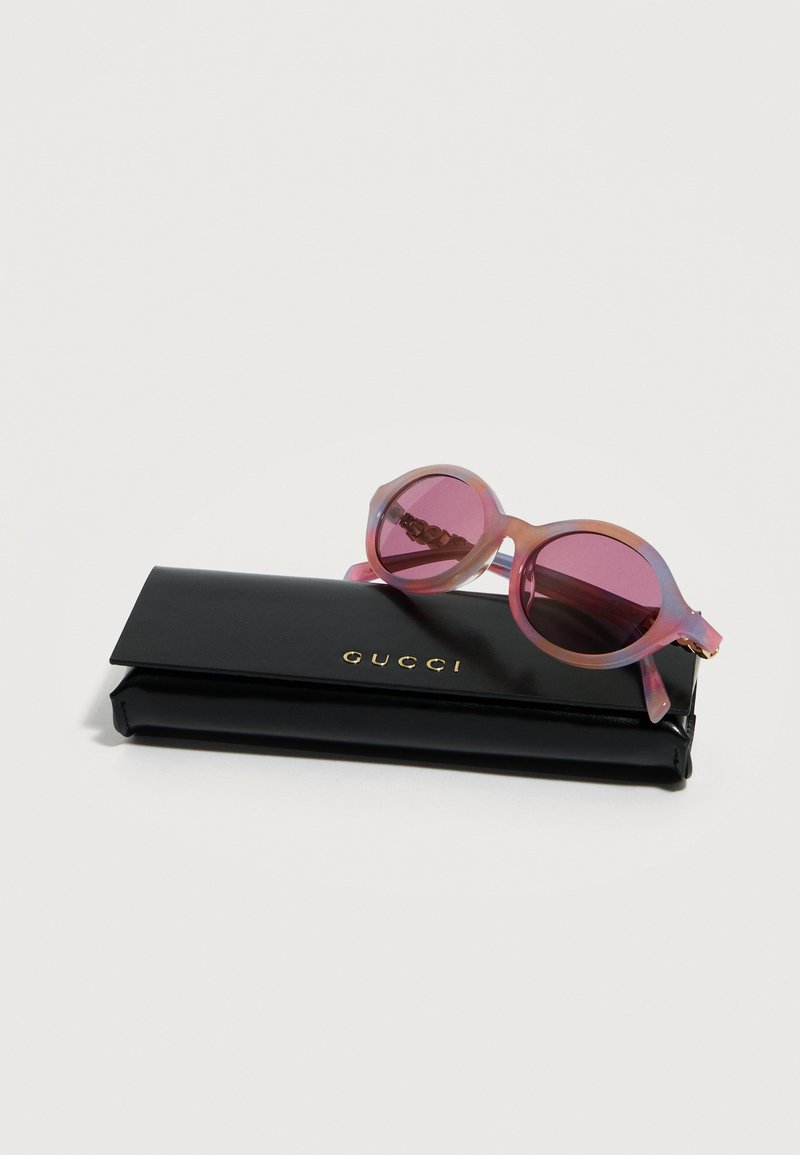 Lunettes de soleil rondes teintées de rose reposant sur un étui Gucci noir avec des lettres dorées sur un fond blanc.