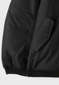 Gros plan sur la manche d'une veste noire montrant une poche à rabat et un poignet élastique côtelé sur un tissu lisse.