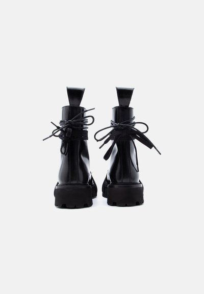 Ζεύγος μαύρων γυαλιστερών δερμάτινων μποτών αστραγάλου με χοντρές σόλες και κορδόνια δεμένα σε φιόγκους, που φαίνονται από πίσω με φόντο λευκό.