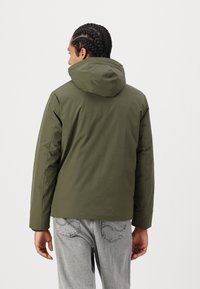 Oliven grønn hettejakke med en glatt, matt finish og glidelås foran. Den har en avslappet passform og er brukt over lysegrå jeans.