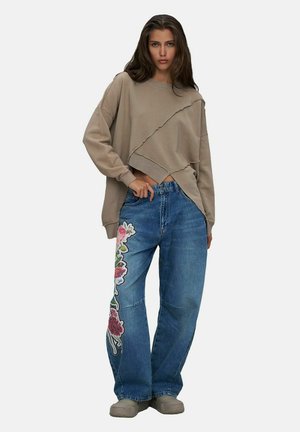 Jeune femme portant un sweat-shirt beige oversize et un jean bleu avec une broderie florale sur la jambe gauche, debout avec une main sur la taille.
