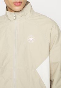 Primer plano de una persona que lleva una chaqueta beige con cremallera y un logo blanco de Converse All Star en el pecho, con paneles blancos contrastantes.