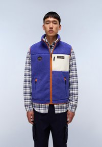 Flis veste u ljubičastoj boji s bež naglascima, sadrži zip džep, narančaste detalje i okrugli logo. Nosena preko košulje s kariranim dezenom.