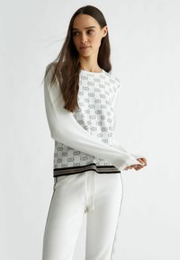 Maglione bianco lavorato a maglia con un motivo ripetuto, polsini a coste e orlo a strisce nere e oro, abbinato a pantaloni bianchi coordinati.