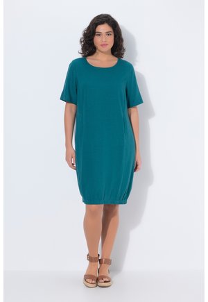 Kvinde med krøllet hår iført en teal knælængde kjole med korte ærmer og brune sandaler med ankelrem, stående mod en hvid baggrund.