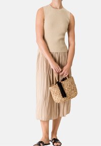 Beige žebrovaná bezrukávová halenka s plisovanou midi sukní, držící tkanou slaměnou tašku s černými třásněmi, doplněná černými sandály.
