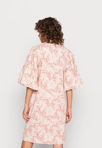 Nainen, jolla on kihara hius, pukeutuneena polvipituiseen, vaaleanpunaiseen mekkoon, jossa on punaisia kukkakuvioita ja leveät puffihihat, näkyy selkäpuolelta.