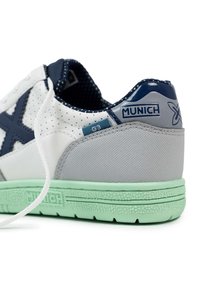 Zapatilla blanca y gris con acentos en azul marino, parte superior perforada, suela de goma verde y logo decorativo en la parte trasera. Diseño con cordones.