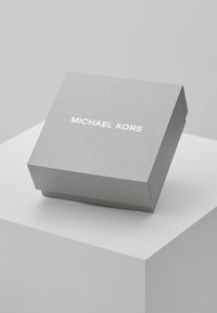 Caja de regalo texturizada en gris con una tapa ligeramente elevada. En la parte frontal presenta "MICHAEL KORS" en letras blancas en negrita. Diseño simple y moderno.