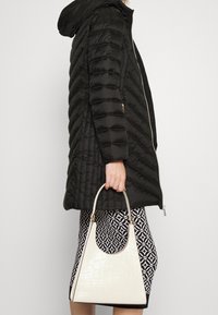 Schwarze Steppjacke mit gestepptem Muster, einem kurzen Reißverschluss und einer Kapuze, kombiniert mit einem gemusterten Rock und einer weißen Handtasche mit Krokodilleder-Optik.