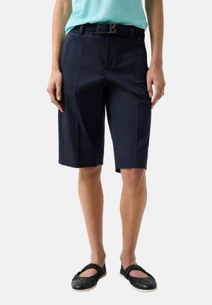 Person iført knælange marineblå skræddersyede shorts med bælte, lyseblå top og sorte slip-on sko, stående mod en ensfarvet baggrund.