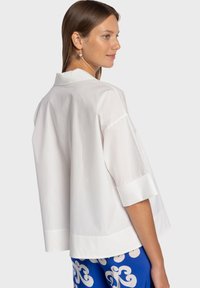 Chemise blanche oversize avec col, manches courtes et détails aux poignets. Texture lisse, design minimaliste, portée avec un pantalon bleu à motifs.