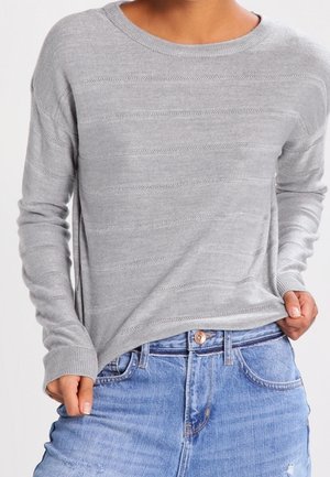 Personne portant un pull à manches longues gris clair avec un motif tricoté horizontal subtil et un jean bleu en denim, les mains tirant légèrement sur le pull.