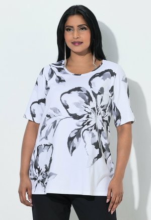 Frau mit langen schwarzen Haaren, die ein weißes kurzärmeliges Hemd mit großen schwarzen und grauen Blumenmustern und schwarze Hose trägt.