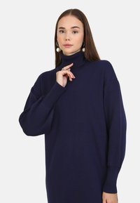 Maglione a collo alto blu navy con vestibilità ampia, polsini a coste e una superficie liscia. Dettagli notevoli includono spalle scese e maniche lunghe.