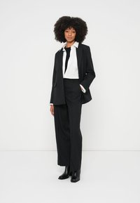 Zwart op maat gemaakt pak met een lang jasje, wijde broekspijpen en een wit overhemd. Voorzien van een zwarte stropdas en enkellaarzen. Soepele textuur en scherp ontwerp.