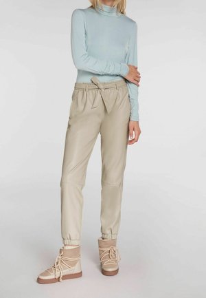 Femme portant un haut bleu clair à manches longues, un pantalon beige taille haute avec ceinture nouée, et des bottines beige à lacets épais, debout devant un fond blanc.