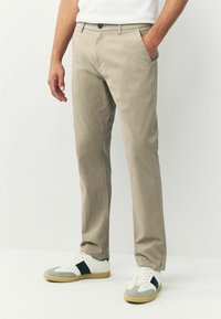 Beige pamut nadrág, slim fit szabással, gombos záródással, két oldalsó zsebből és sima textúrával. Fehér és szürke sportcipővel viselve.